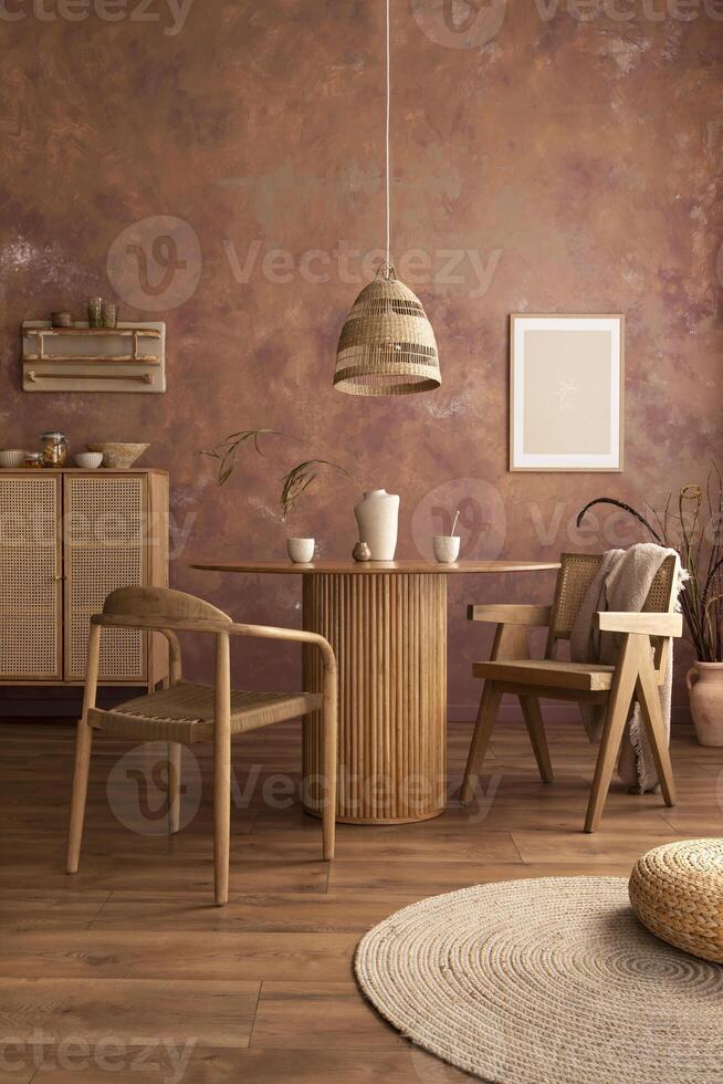 Stylish boho vintage dining room interior with poster mock up. Round table with rattan armchair, round carpet with pouf, vase with dried flowers and cup. Brown wall and rattan lamp. Mock up poster. photo