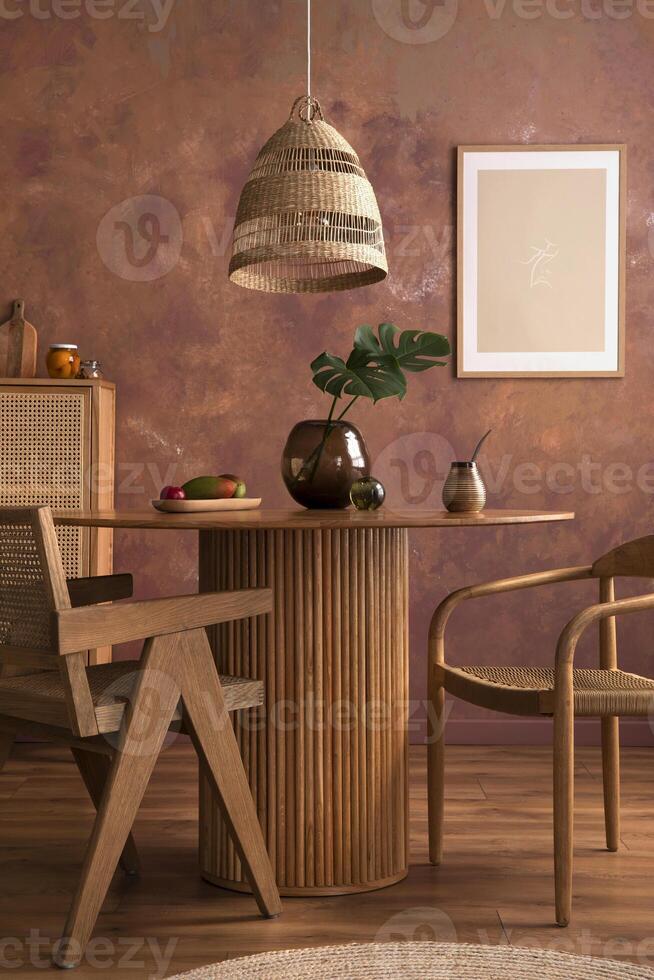Stylish boho vintage dining room interior with poster mock up. Round table with rattan chair, vase with dried flowers and rattan armchair. Brown wall and rattan lamp. Mock up poster. Template. photo