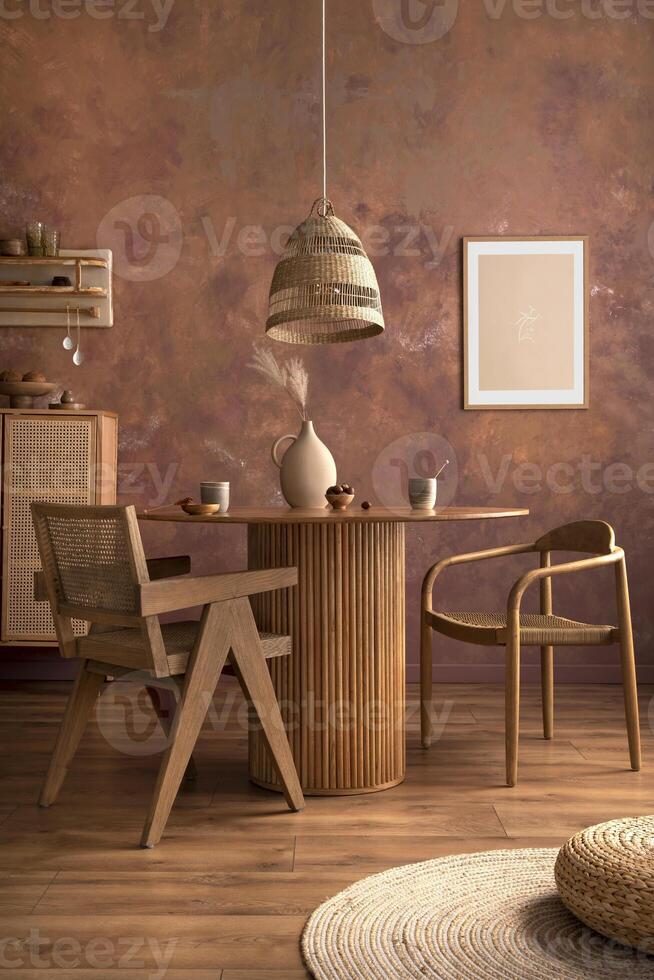 Stylish boho vintage dining room interior with poster mock up. Round table with rattan chair, vase with dried flowers. Brown wall and rattan lamp. Mock up poster. Template. photo