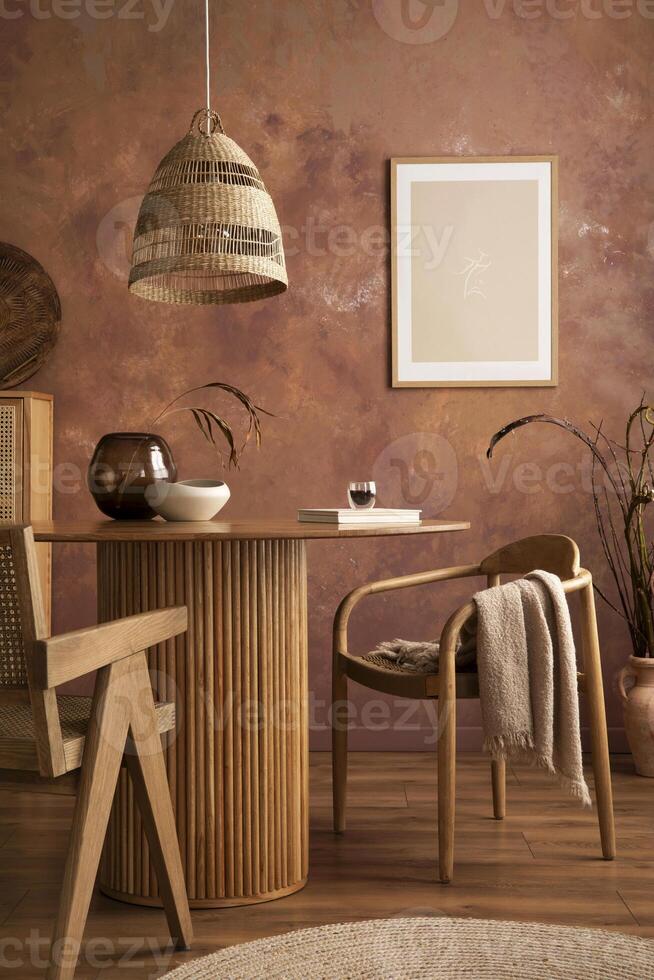 Stylish boho vintage dining room interior with poster mock up. Round table with rattan chair, vase with dried flowers. Brown wall and rattan lamp. Mock up poster. Template. photo