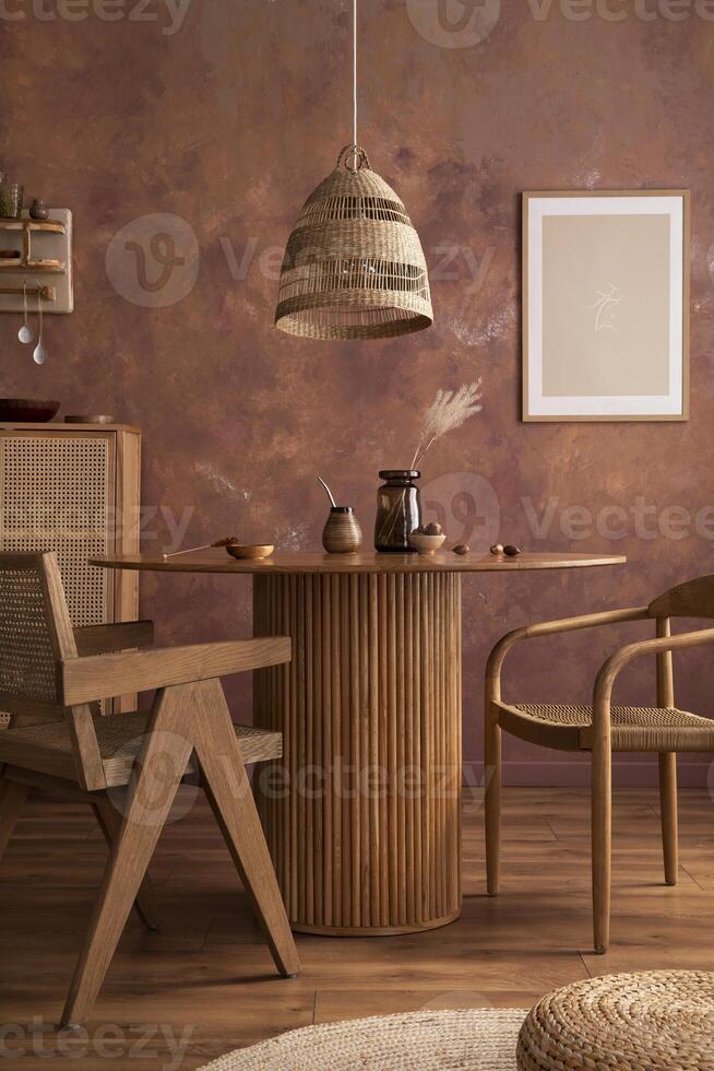 Stylish boho vintage dining room interior with poster mock up. Round table with rattan chair, vase with dried flowers. Brown wall and rattan lamp. Mock up poster. Template. photo