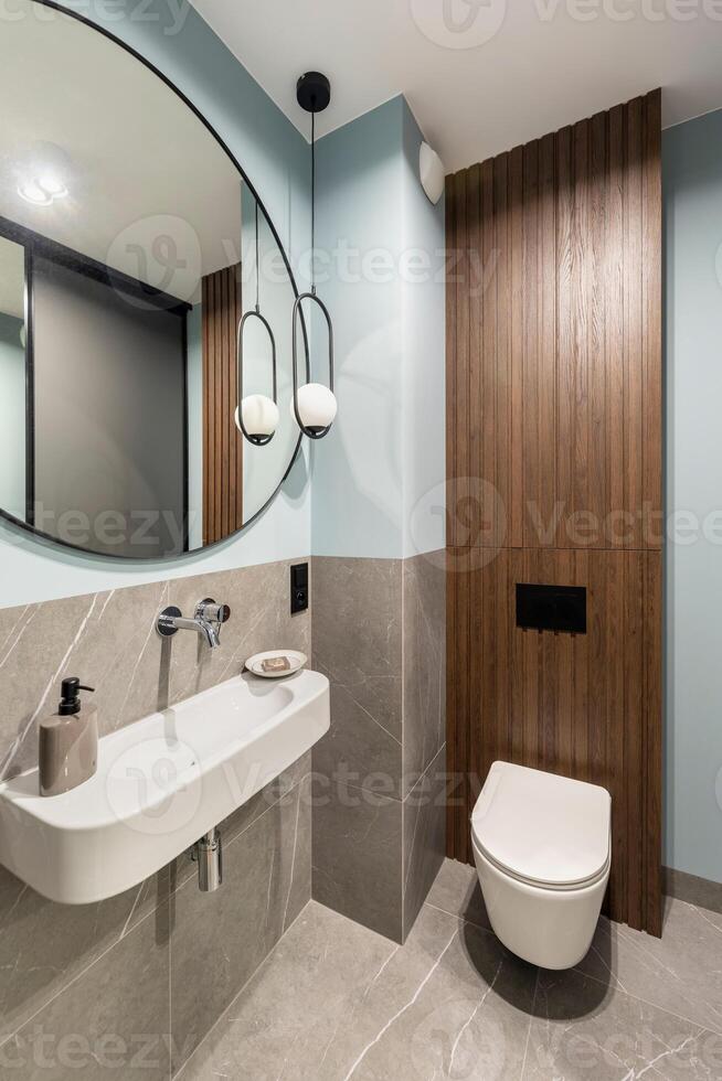 Modern bright bathroom with white toilet seat on wooden lamela wall. Round mirror with black frame. photo