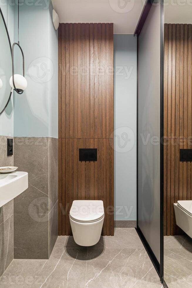 Modern bright bathroom with white toilet seat on wooden lamela wall. Mirror on grey wall. photo