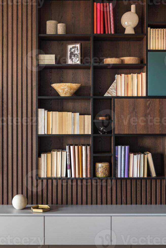 Modern bookcase in lamells wall. Grey chest of drawers. Bookcase a lot of books and accessories. Herringbone wooden parquet. Cat in open spaces. photo