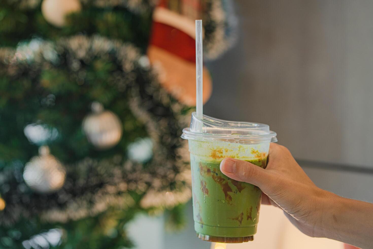hand of woman holding a take-out iced matcha caramel with blurred christmas tree background at cafe, isolate concept. photo