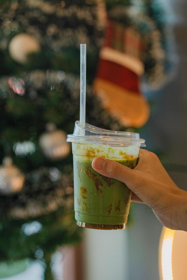 hand of woman holding a take-out iced matcha latte with blurred christmas tree background at cafe photo