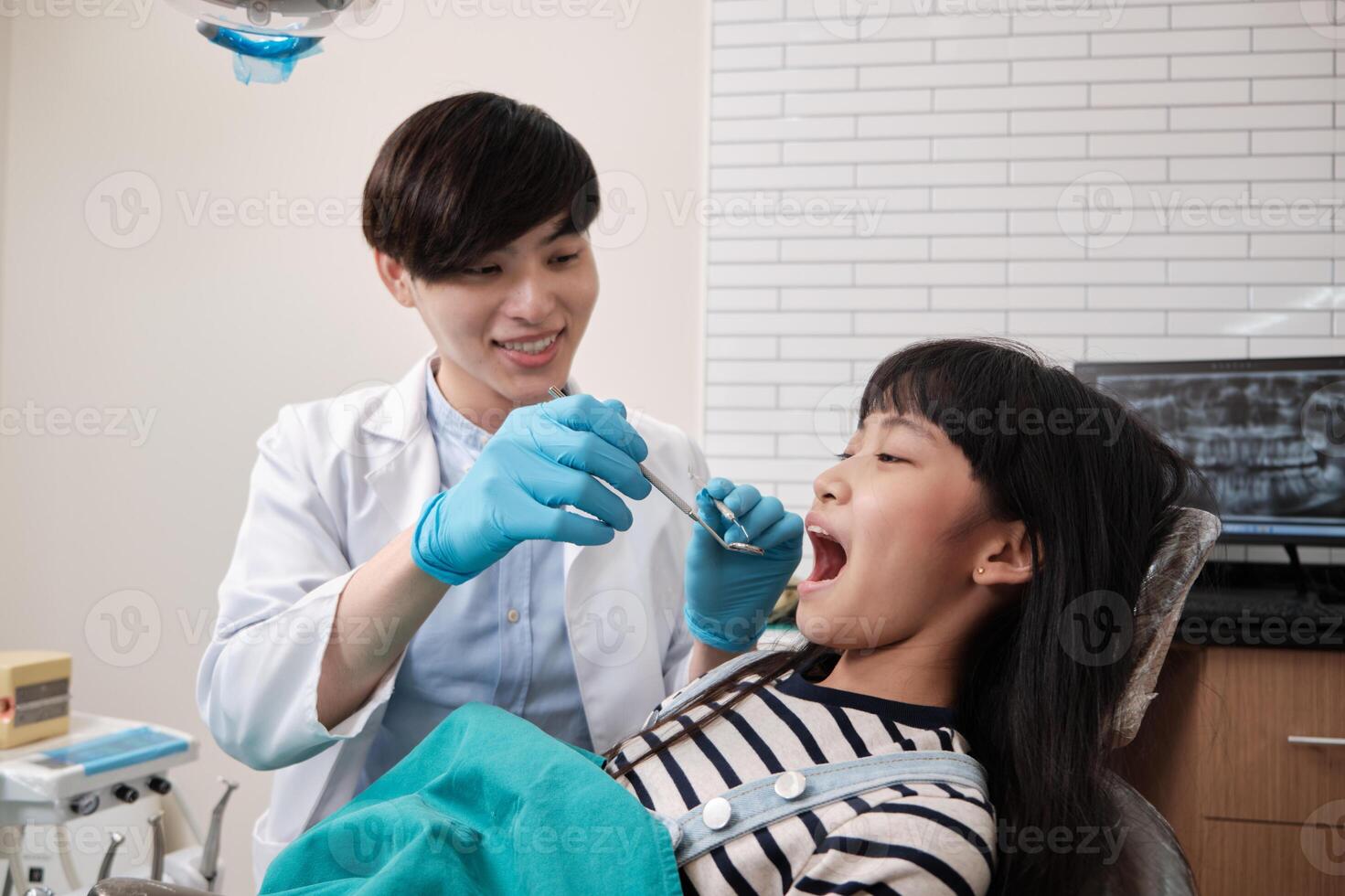 A young Asian male pediatric dentist checks and examines a little Thai girl's teeth in dental clinic, well-being mouth oral hygiene, and professional orthodontic healthcare work in a kid hospital. photo