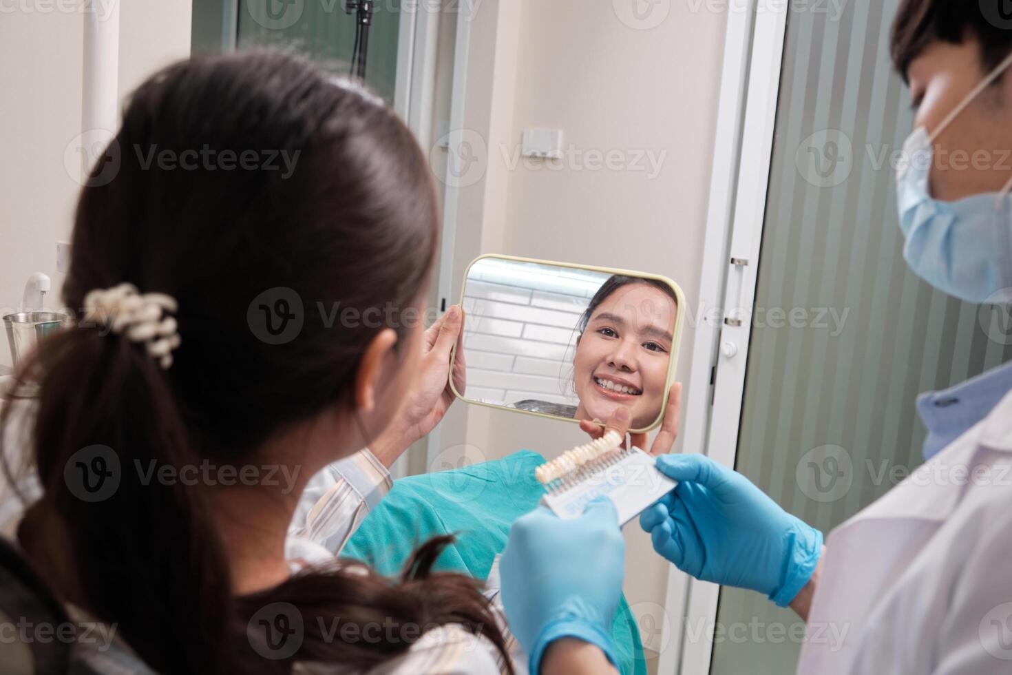 Asian male dentist explains color shades of tooth implant whitening to young female patient, look at mirror in dental clinic, and do professional mouth hygiene healthcare work in a doctor's hospital. photo