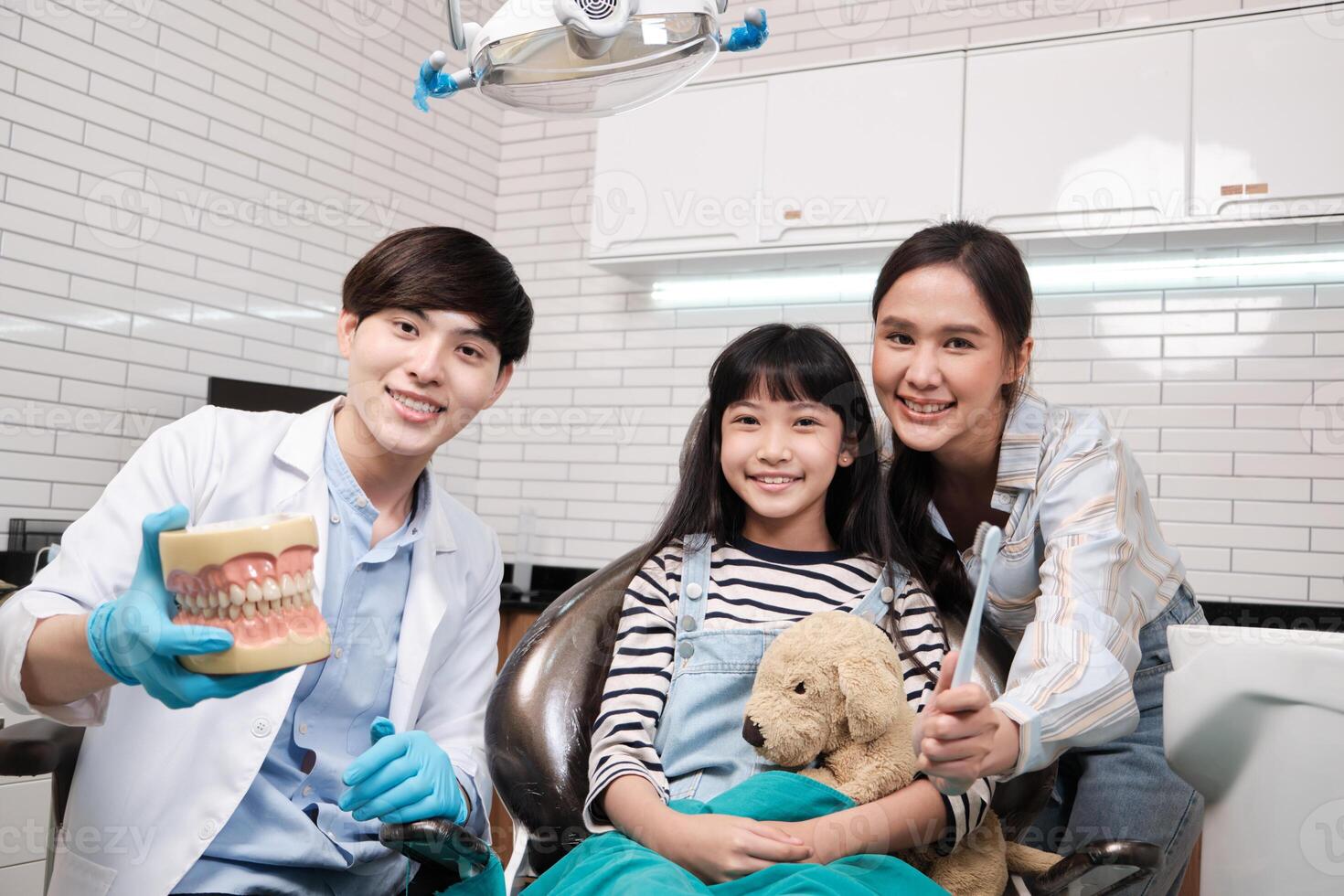 Portrait of Asian male pediatric dentist, mother, and a girl patient smiling and looking at camera in dental clinic, mouth hygiene, and professional orthodontic healthcare work in a kid hospital. photo