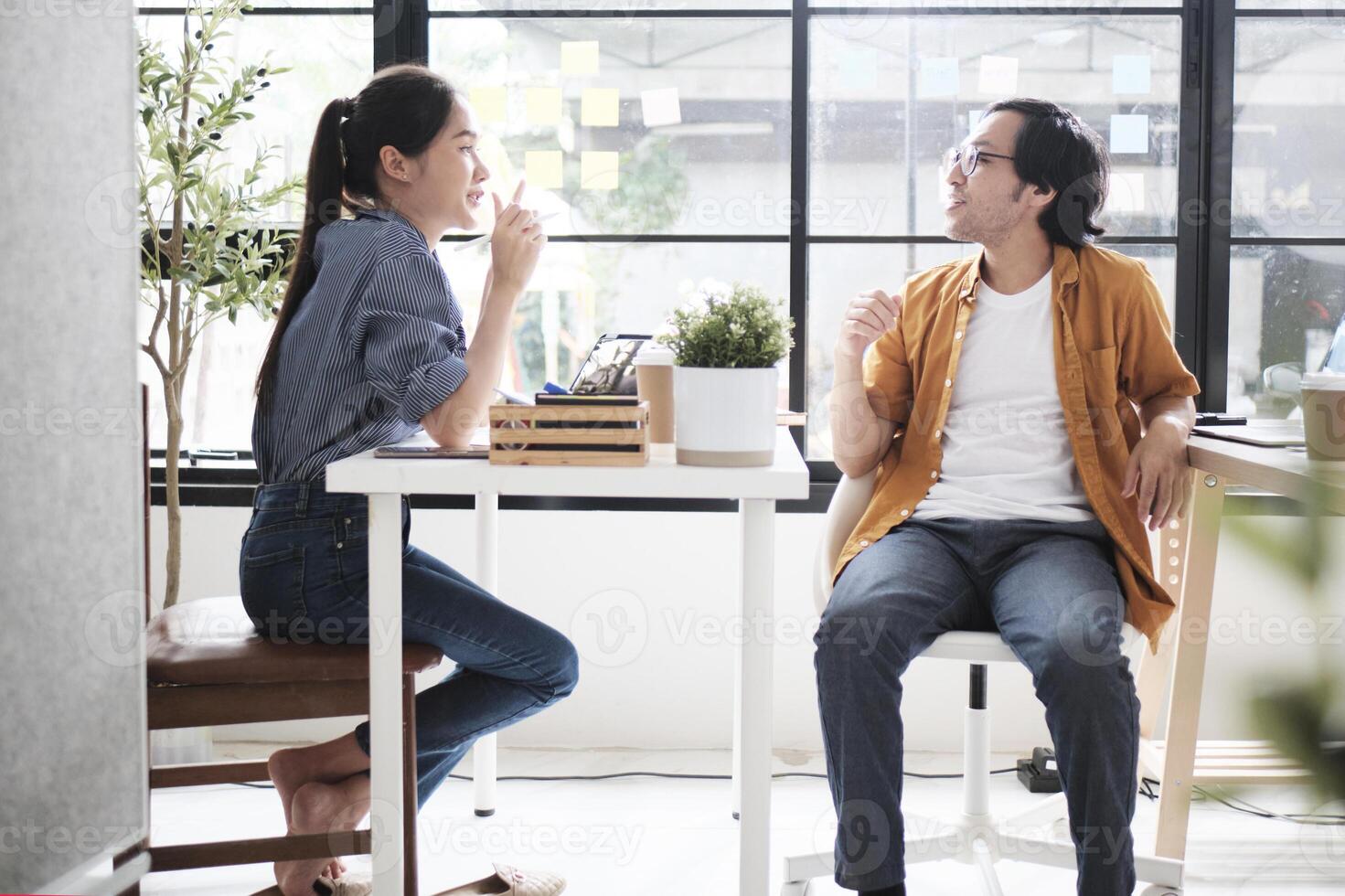 Two Asian workers sitting in chairs at working desks, collaborating and talking at daylight windows in casual small business office, online business job coworkers, positive relationship colleagues. photo