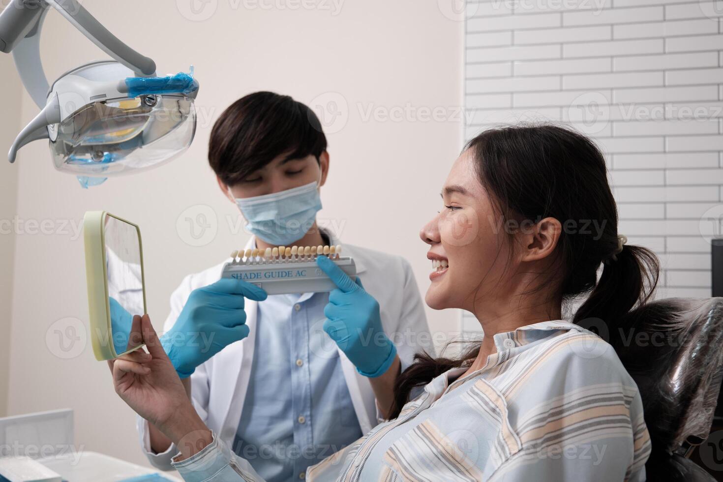 Asian male dentist explains color shades of tooth implant whitening to young female patient, look at mirror in dental clinic, and do professional mouth hygiene healthcare work in a doctor's hospital. photo