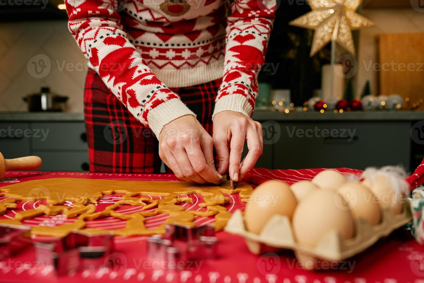 Woman in festive sweater using cookie cutters to shape gingerbread dough on red mat. Baking traditional Christmas cookies in home kitchen photo