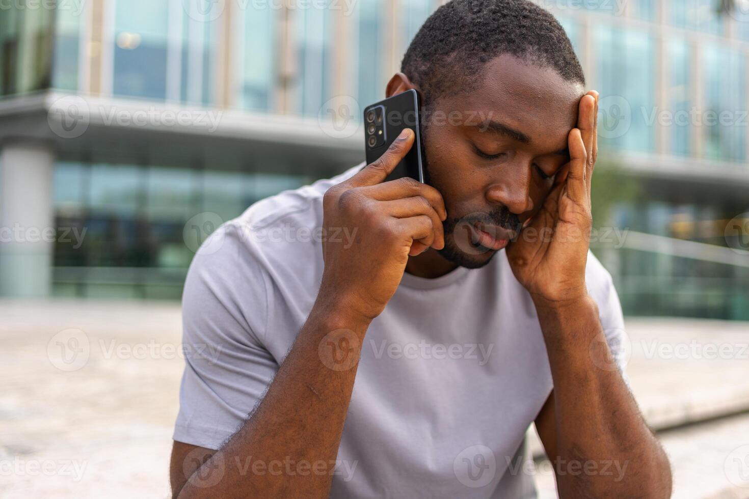 Sad tired lonely disappointed African man alone talking on smartphone on city street outdoor. Unhappy person making answering call by cellphone experiencing grief getting bad news. Stressed man photo