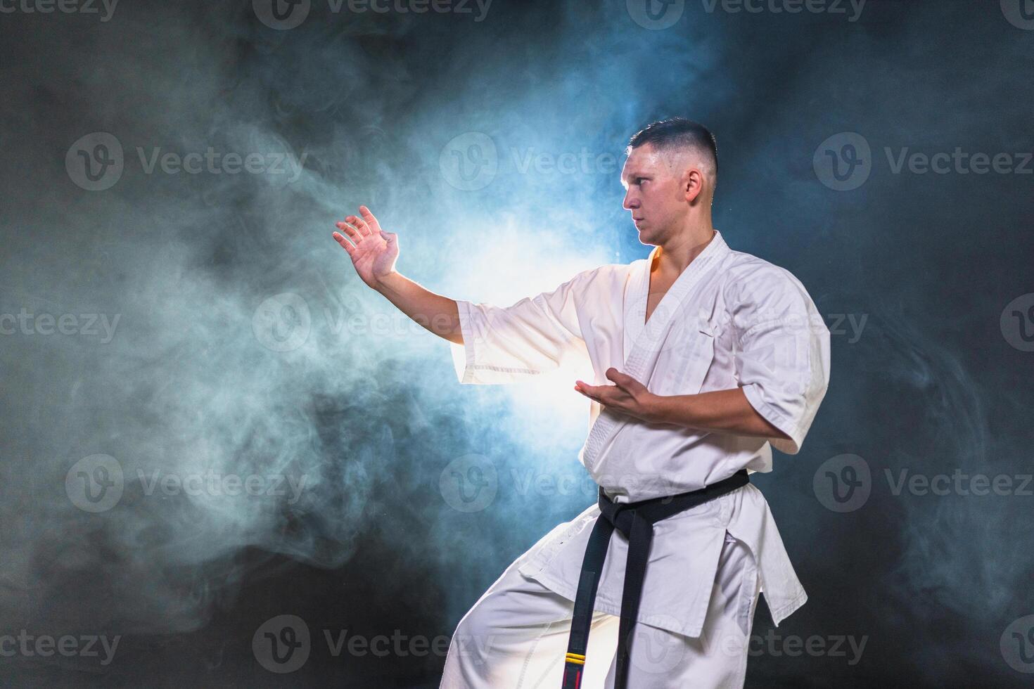 Strong man fighter in white kimono for karate martial arts with black belt preparing for fight training in black background. Man training moves punches kicks practicing shadow fight ready for sparring photo