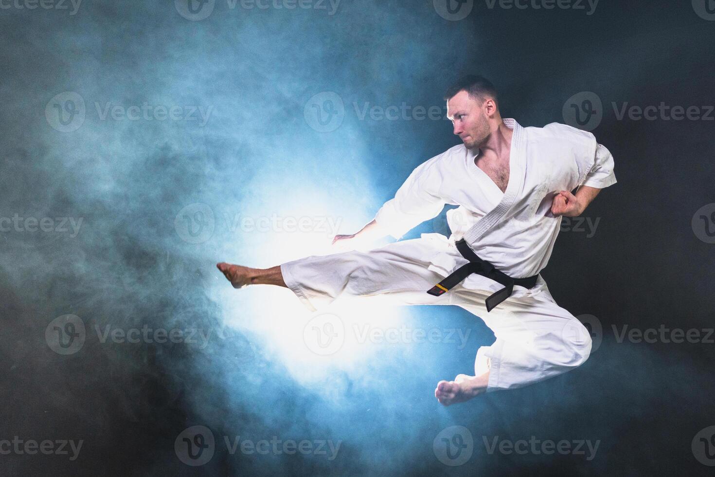 Strong man fighter in white kimono for karate martial arts with black belt jump kicking in black background. Man training moves practicing shadow fight preparing for fight training ready for sparring photo