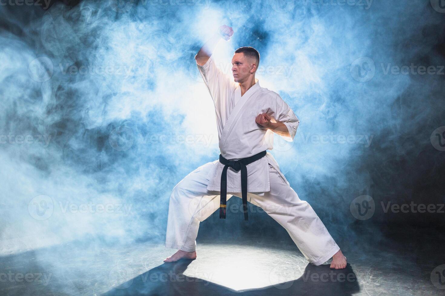 Strong man fighter in white kimono for karate martial arts with black belt preparing for fight training in black background. Man training moves punches kicks practicing shadow fight ready for sparring photo