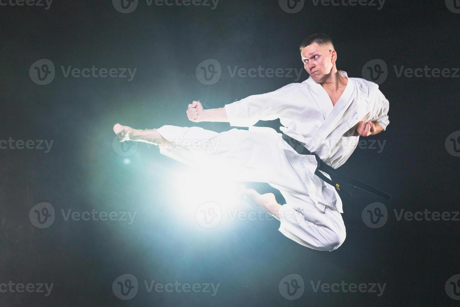Strong man fighter in white kimono for karate martial arts with black belt jump kicking in black background. Man training moves practicing shadow fight preparing for fight training ready for sparring photo