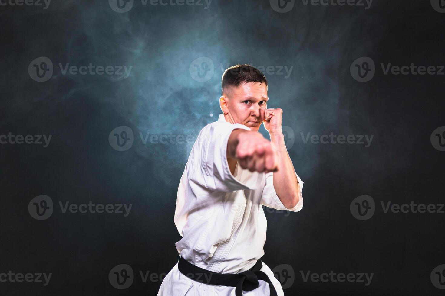 Strong man fighter in white kimono for karate martial arts with black belt preparing for fight training in black background. Man training moves punches kicks practicing shadow fight ready for sparring photo