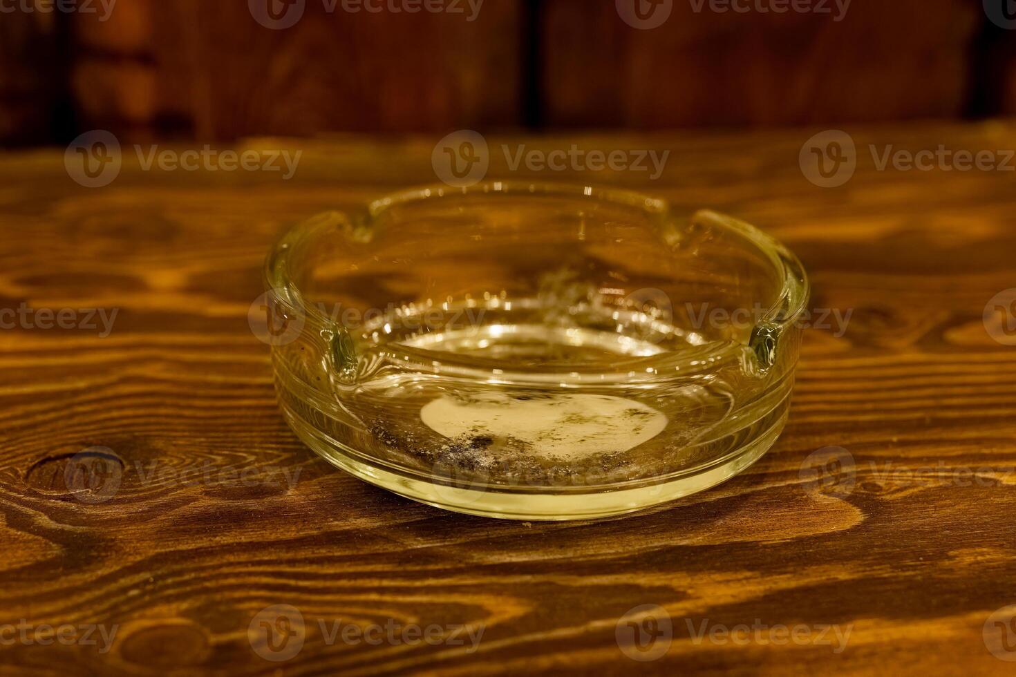 Clear glass ashtray sits on a rustic wooden table with remnants of tobacco at a casual gathering spot in the evening photo