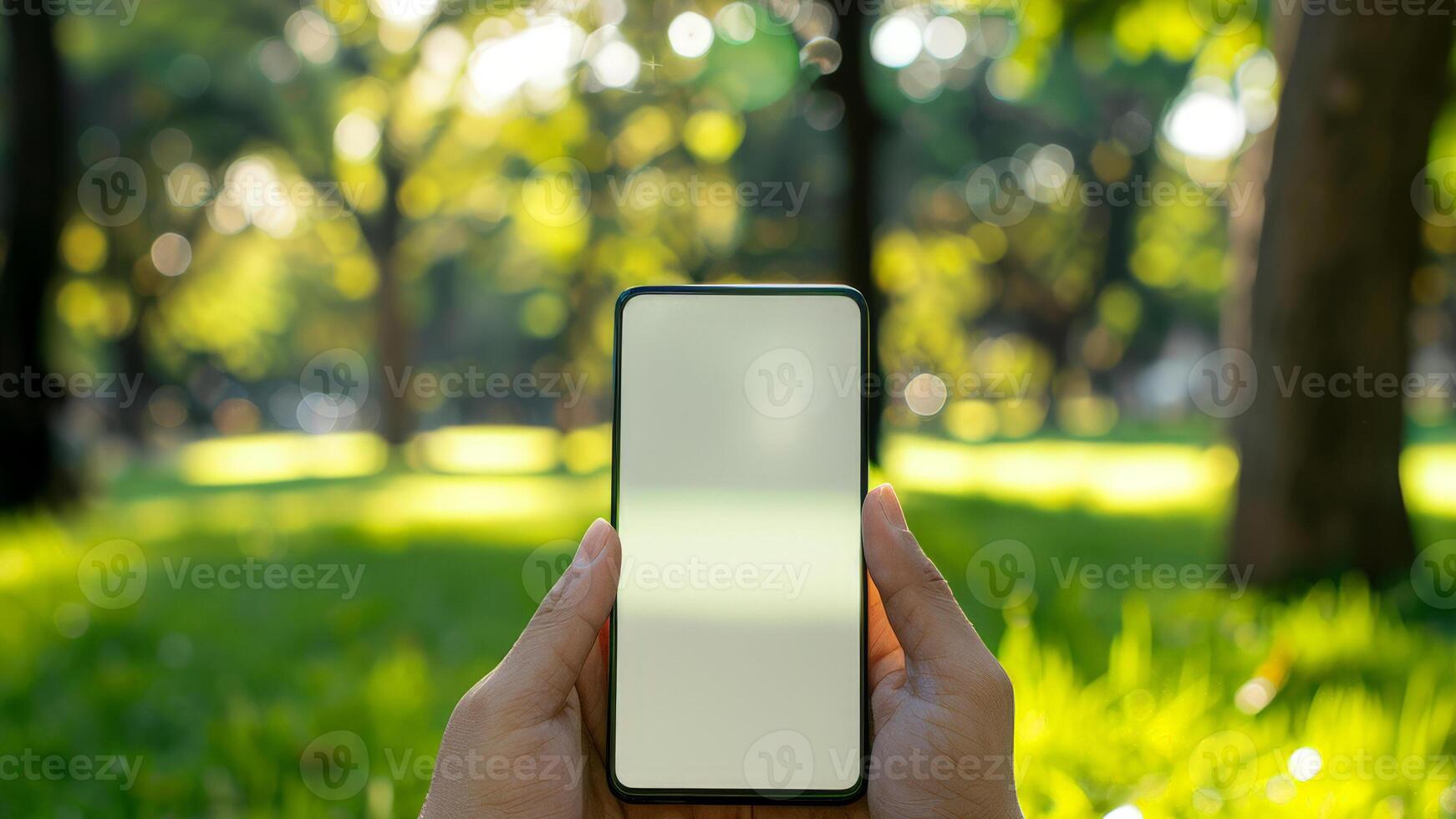 A person gently holds a smartphone featuring an empty screen, surrounded by vibrant greenery and trees in a peaceful park during daylight hours photo