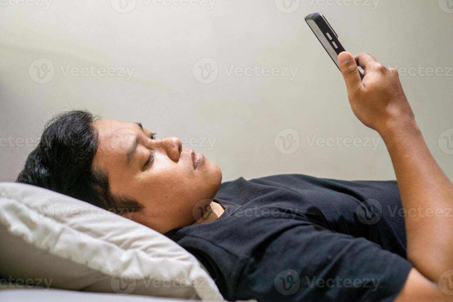 Young Asian man lying on bed while using smart phone. Man using smartphone in bed at night. Bad habit concept. photo