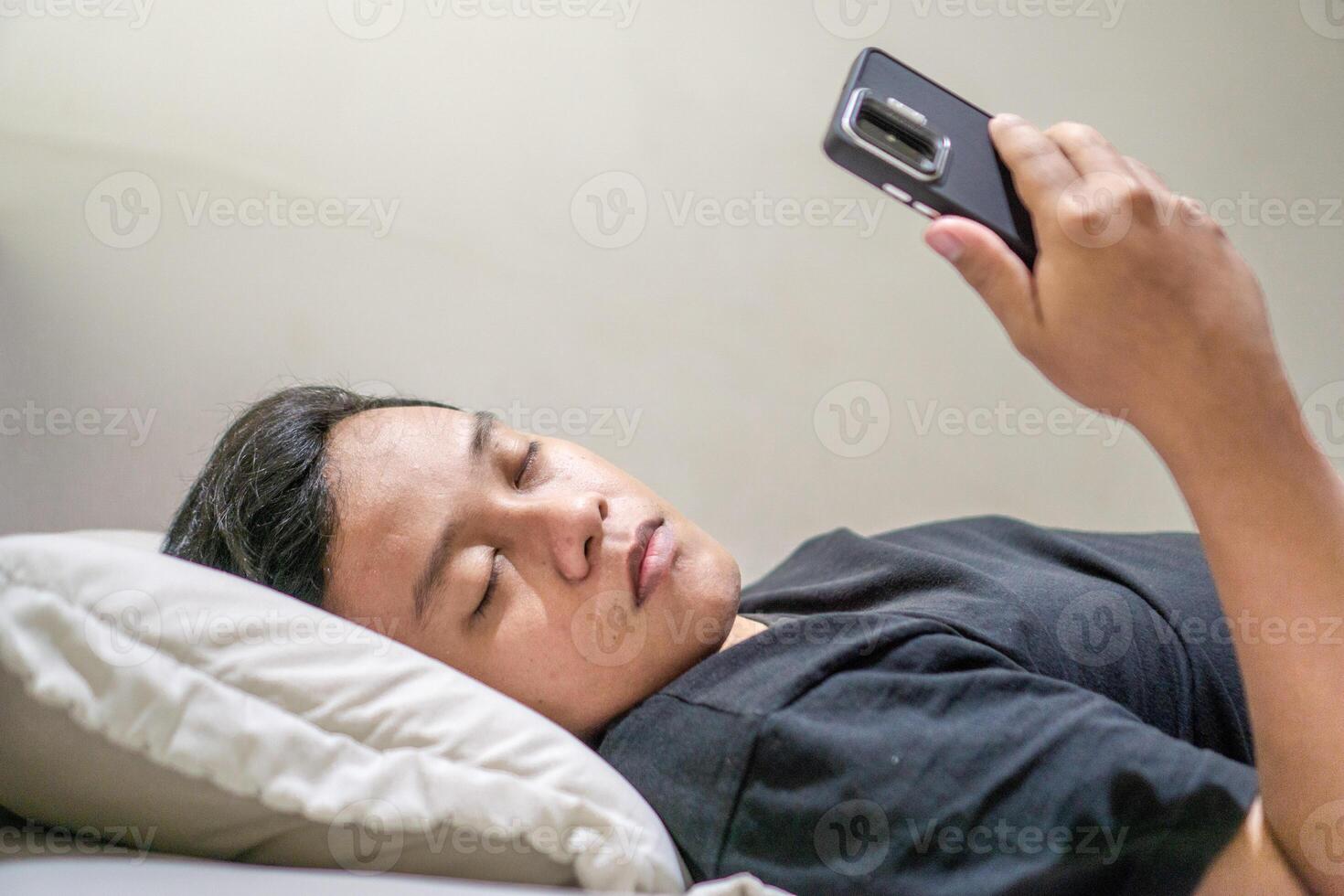 Young Asian man lying on bed while using smart phone. Man using smartphone in bed at night. Bad habit concept. photo