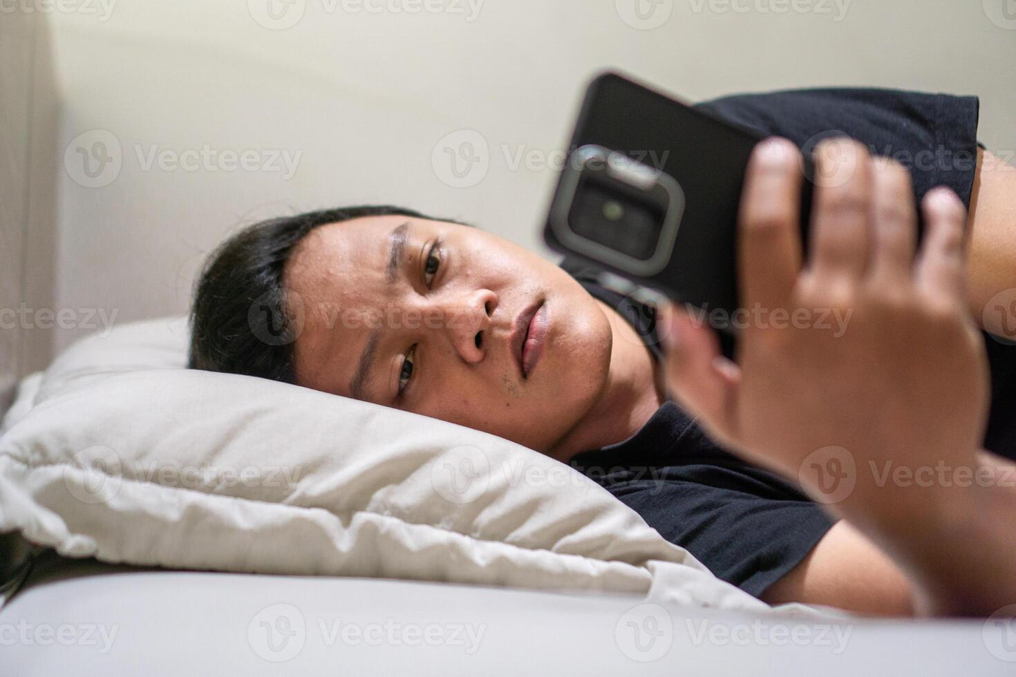 Young Asian man lying on bed while using smart phone. Man using smartphone in bed at night. Bad habit concept. photo
