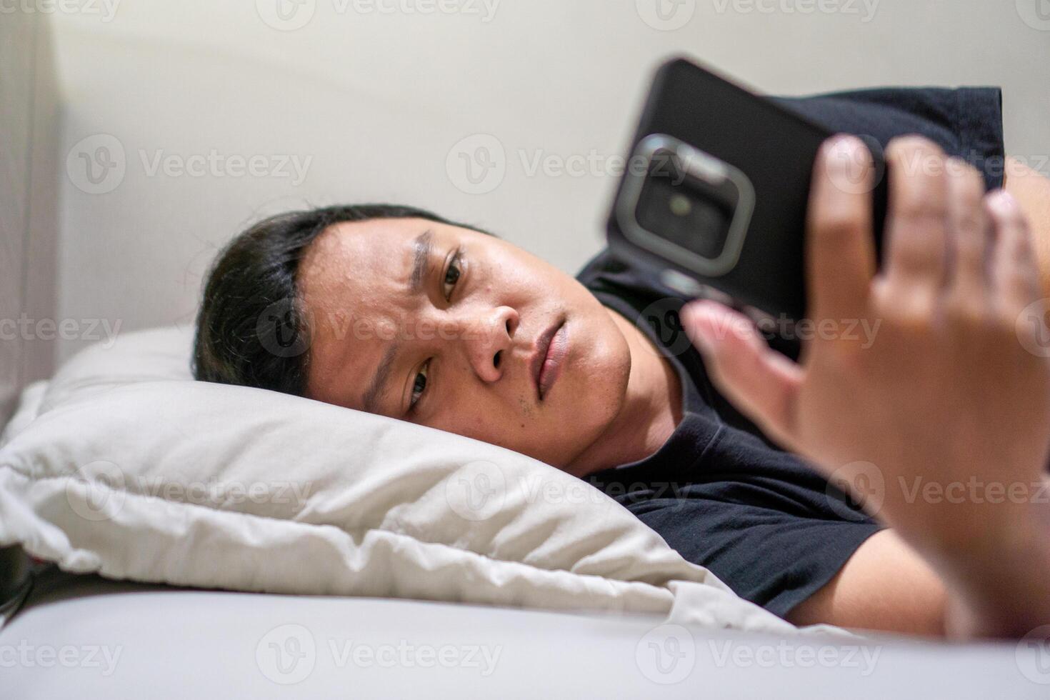 Young Asian man lying on bed while using smart phone. Man using smartphone in bed at night. Bad habit concept. photo