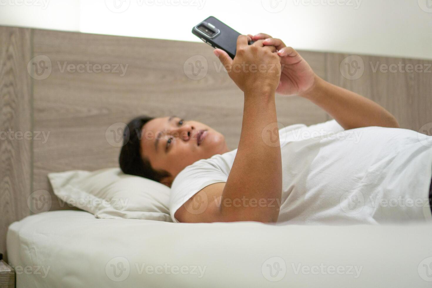 Young Asian man lying on bed while using smart phone. Man using smartphone in bed at night. Bad habit concept. photo