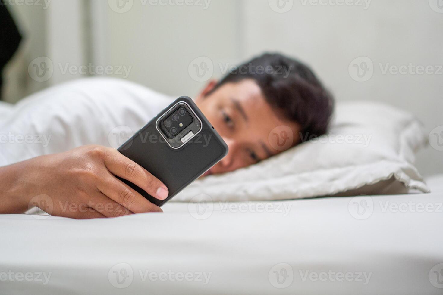 Young Asian man lying on bed while using smart phone. Man using smartphone in bed at night. Bad habit concept. photo