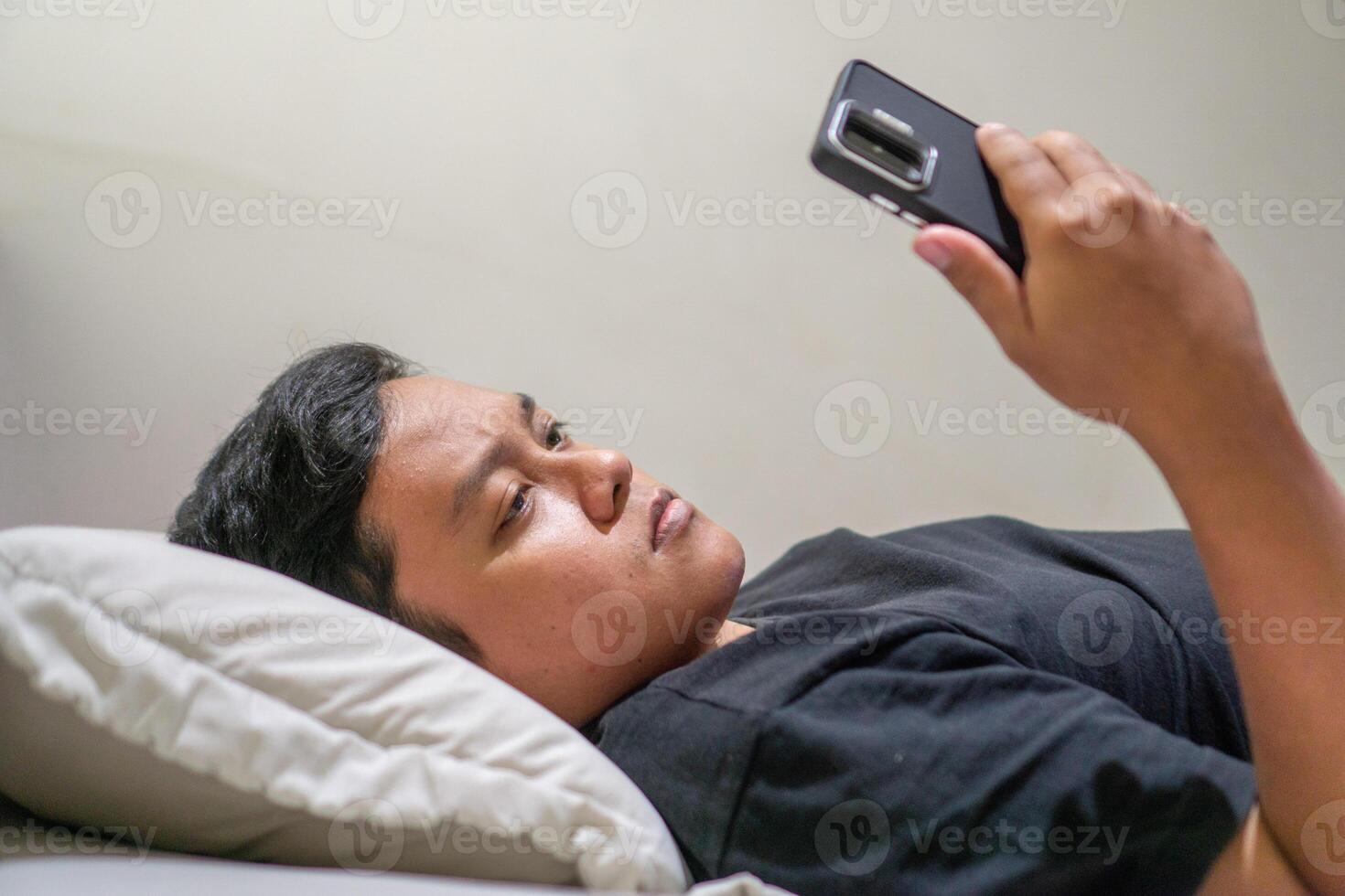Young Asian man lying on bed while using smart phone. Man using smartphone in bed at night. Bad habit concept. photo