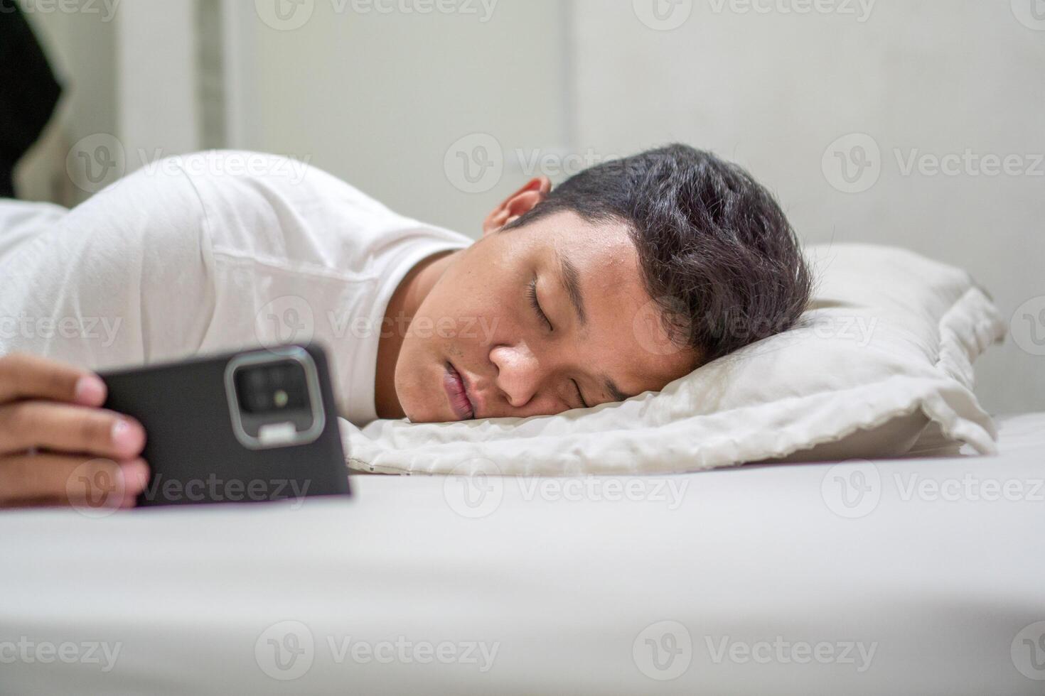 Tired Asian man fall asleep on bed while using mobile phone. Man sleep in bed at night and still holding his phone. Bad habit concept. photo