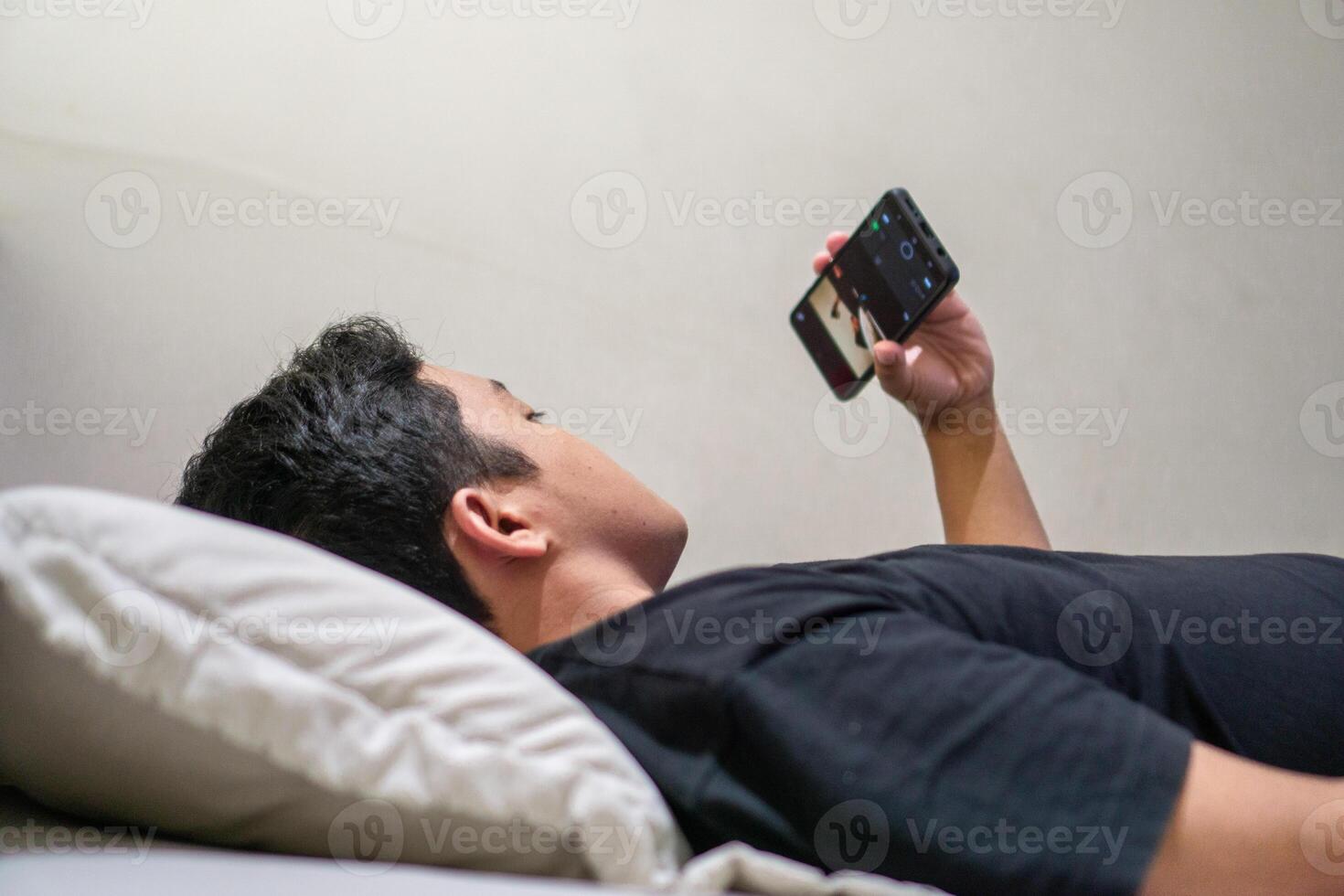Young Asian man lying on bed while using smart phone. Man using smartphone in bed at night. Bad habit concept. photo