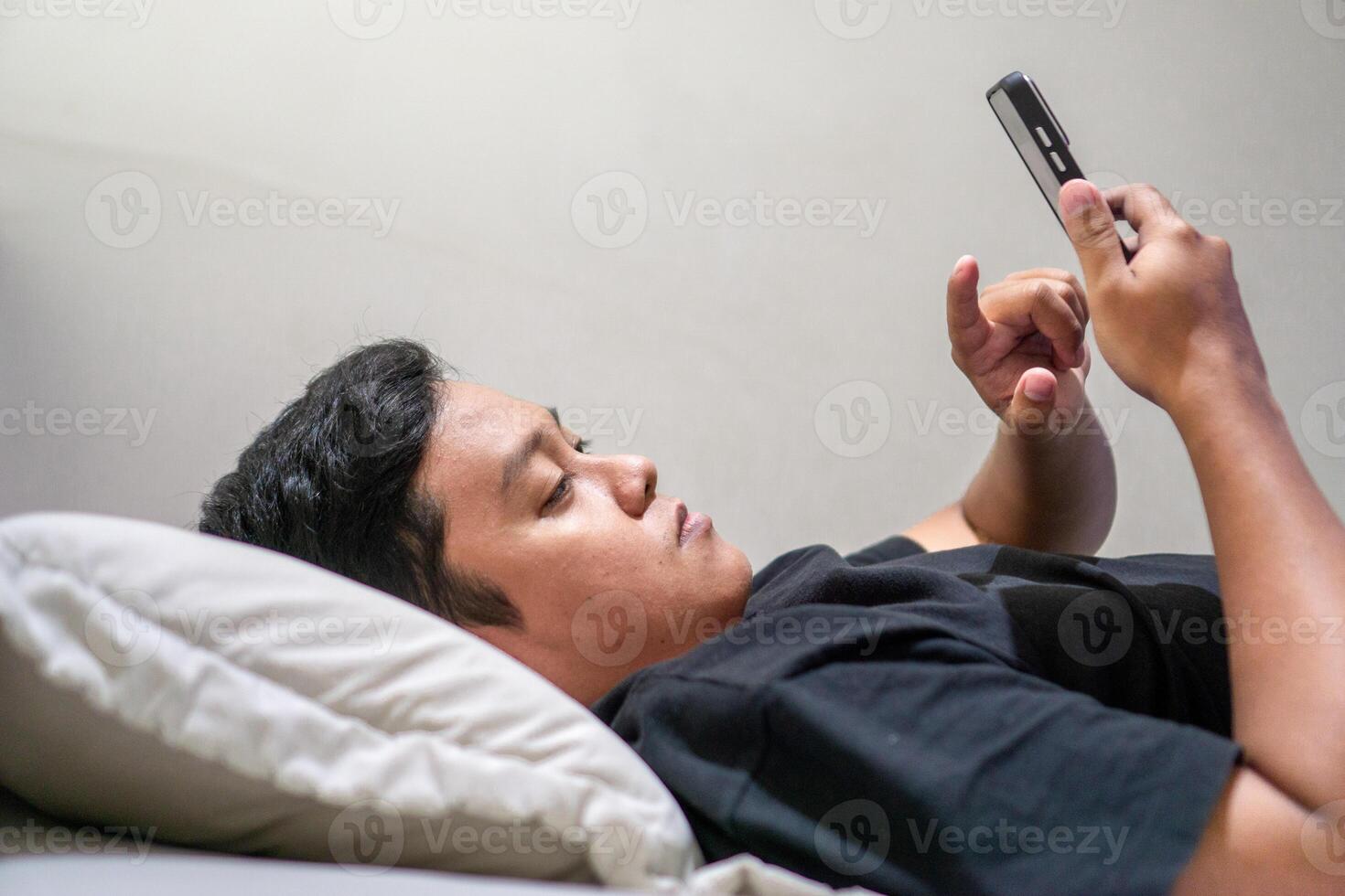 Young Asian man lying on bed while using smart phone. Man using smartphone in bed at night. Bad habit concept. photo