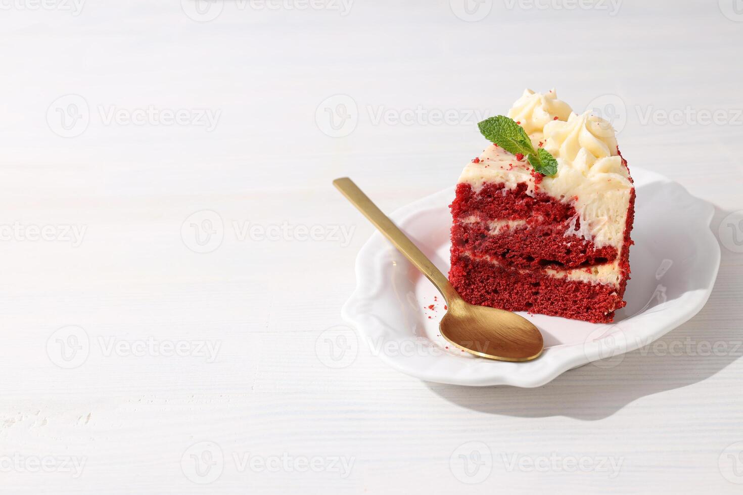 Piece of red velvet cake and spoon on plate on white wooden background, space for text photo