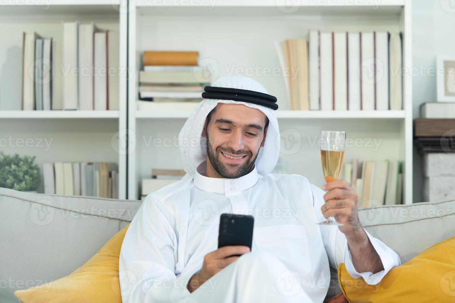 Portrait of a smiling Arabian man holding a glass of champagne and using mobile phone at home photo