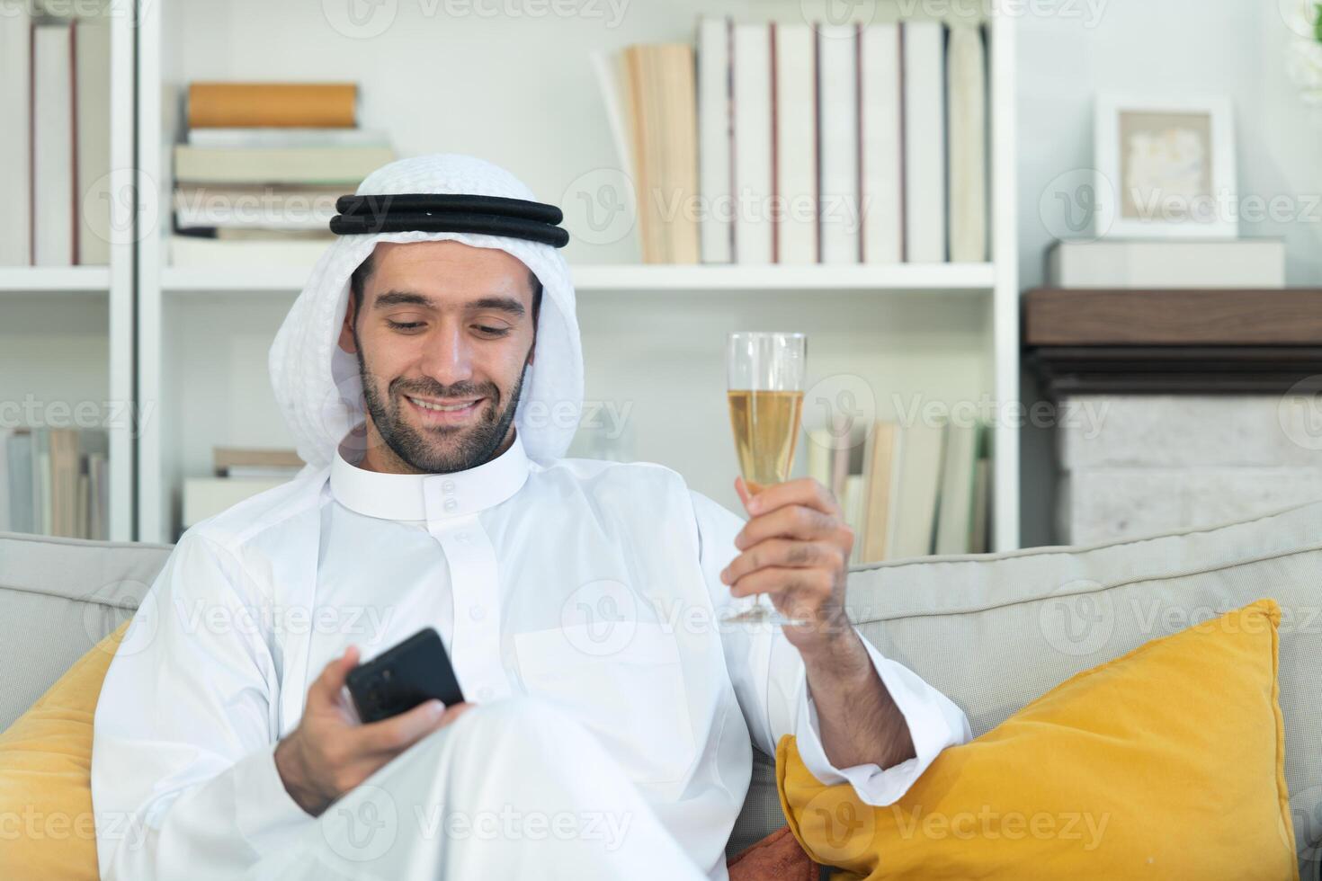 Portrait of a smiling Arabian man holding a glass of champagne and using mobile phone at home photo