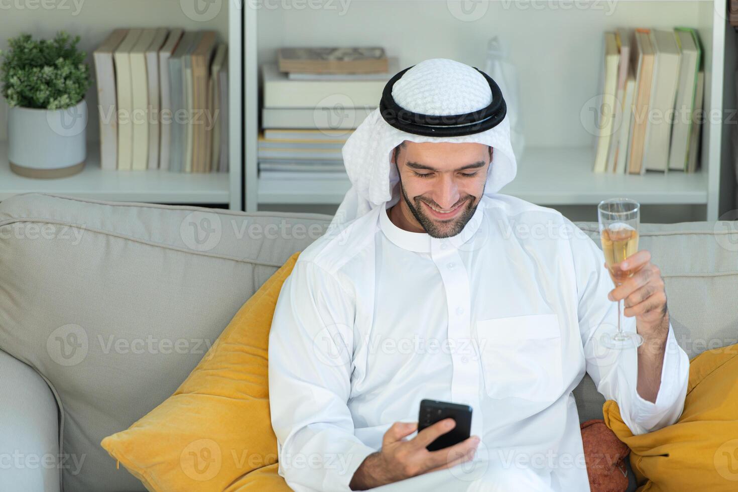 Portrait of a smiling Arabian man holding a glass of champagne and using mobile phone at home photo