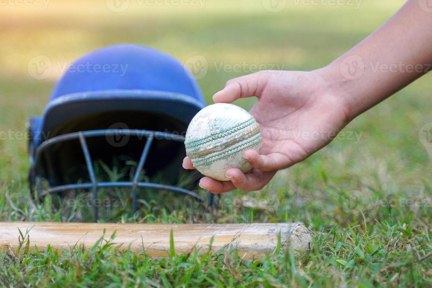 A player's hand picks up a cricket ball from a pile of other cricket equipment on the grass, which includes a cricket helmet, bat and gloves. photo
