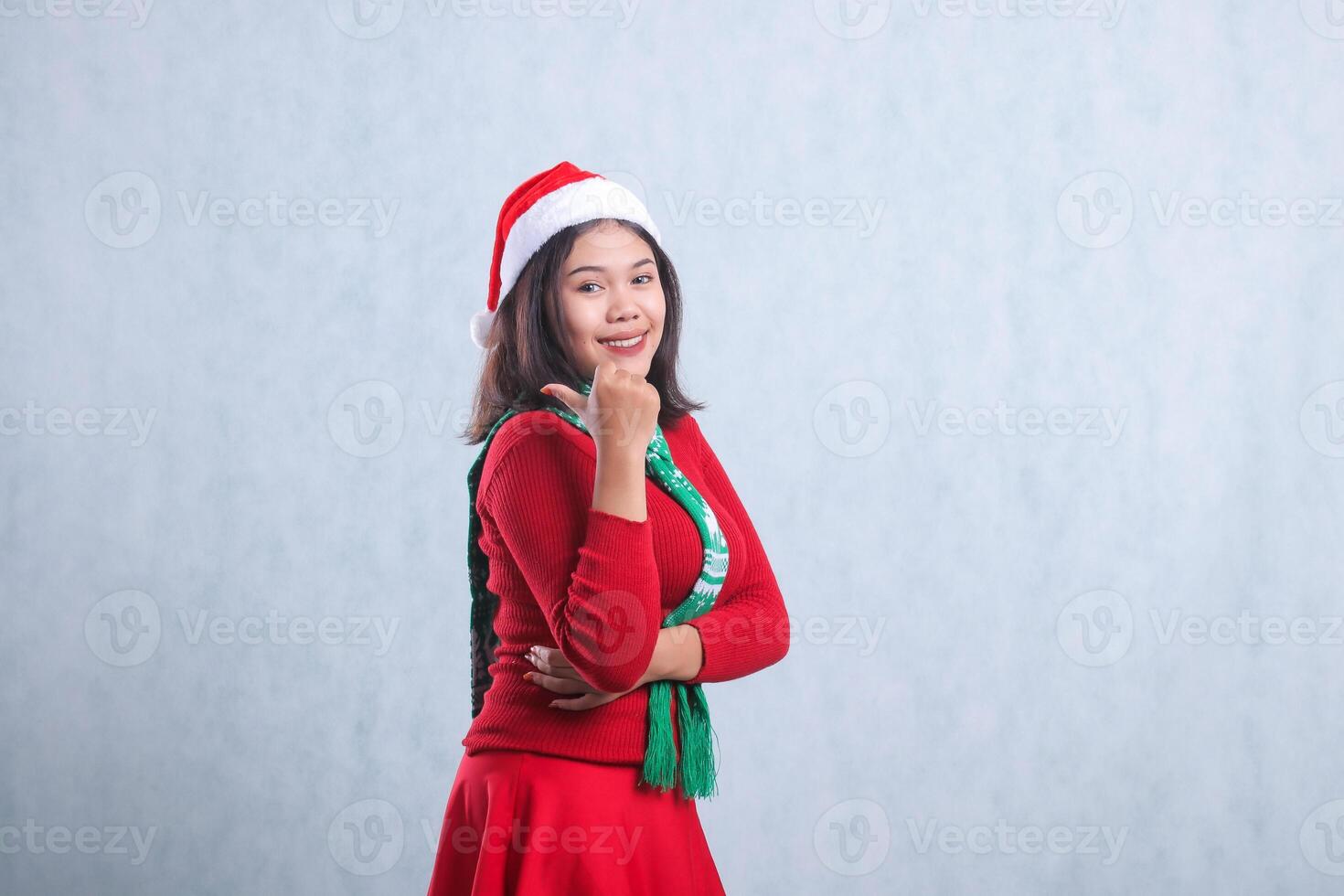 portrait of beautiful girl wearing christmas sweater, santa hat and scarf, slanted left smile to camera hand sign pointing to right, isolated on white background photo
