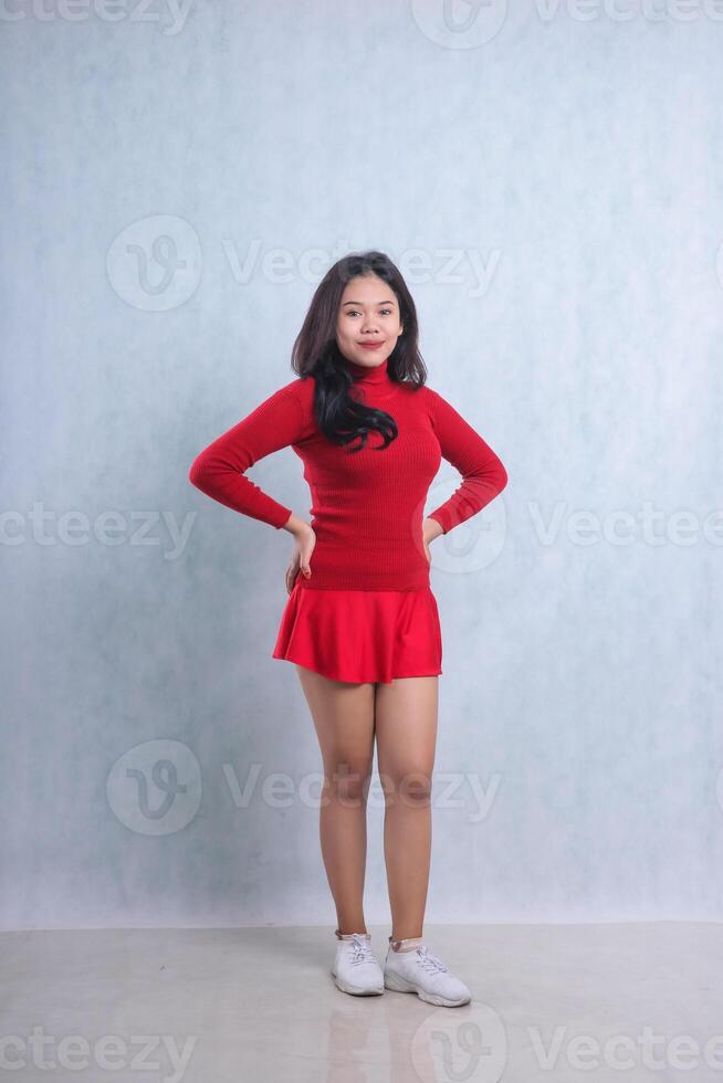 full body gesture of young Asian woman wearing red sweater, standing smiling to camera both hands holding waist, isolated on white background photo