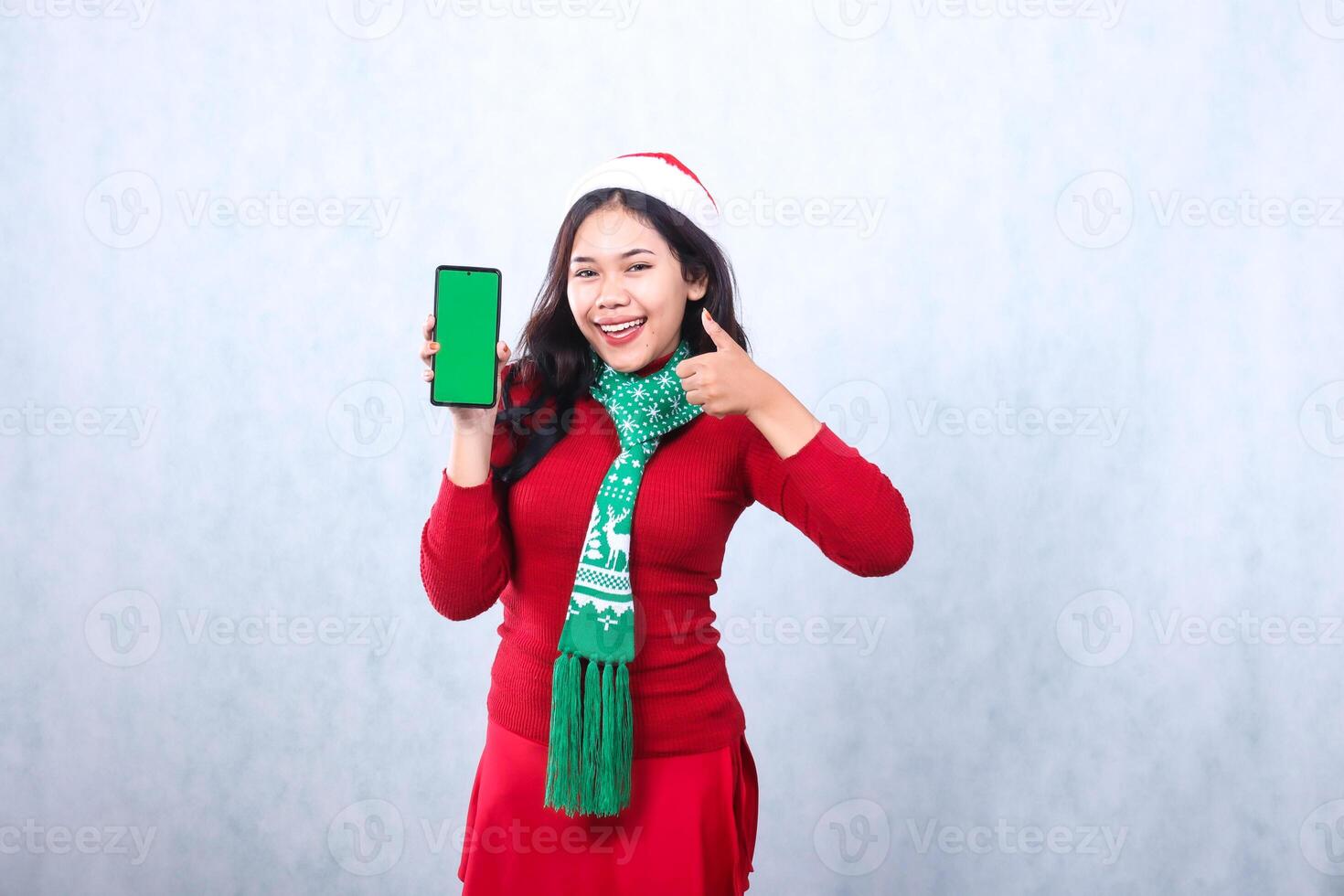 expression of woman wearing red christmas sweater with Santa hat and scarf, cheerful looking at camera holding cellphone mobile phone screen while thumbs up sign, nice, cool, isolated white background photo