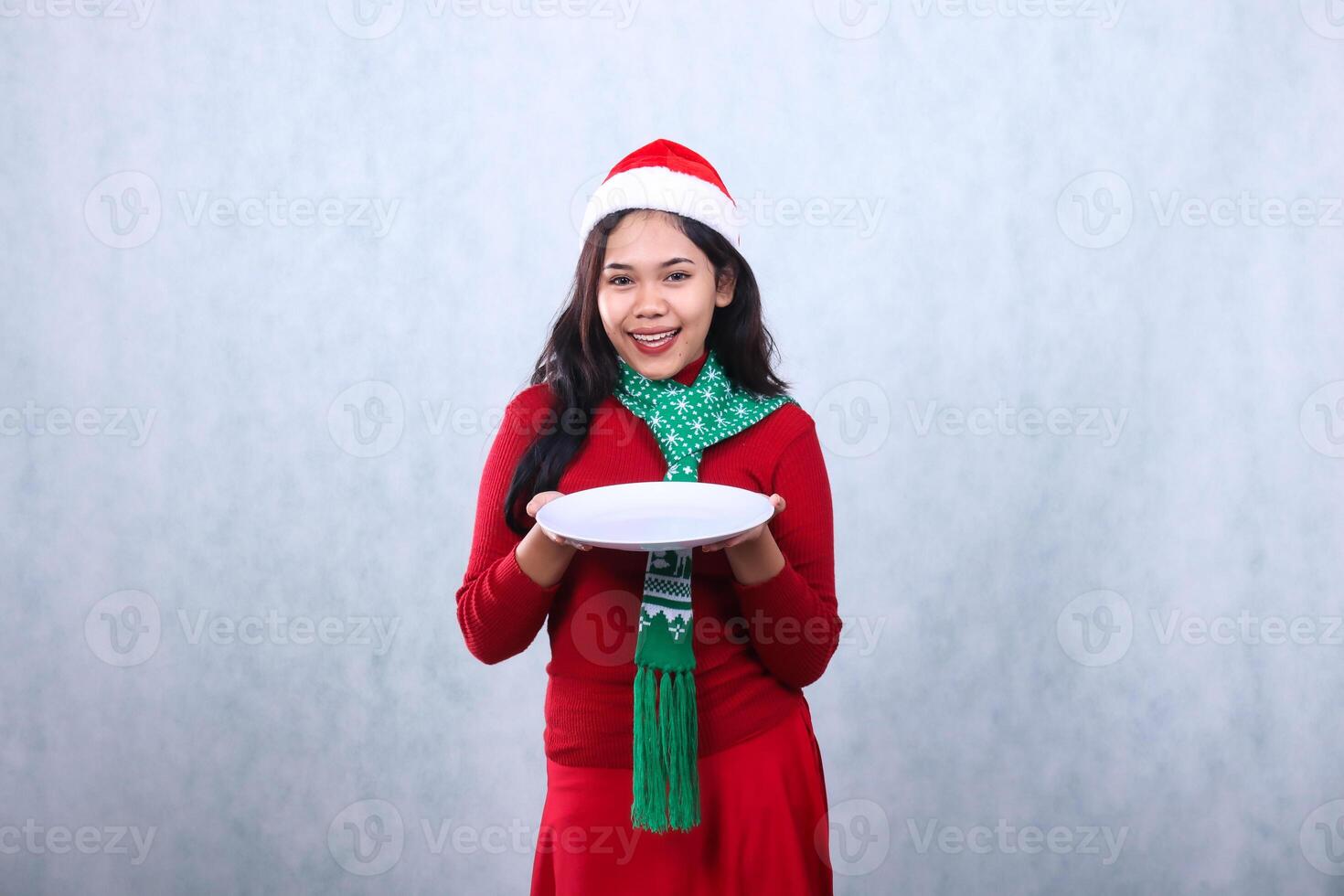 gesture of young Asian woman wearing red sweater with Santa hat and scarf, cheerful to camera both hands carrying white plate, celebrating merry christmas and new year isolated on white background photo