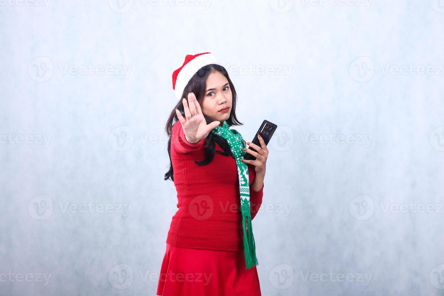 adult asian girl serious to camera hand holding mobile phone and stop sign, wearing red sweater with Santa hat and scarf celebrating merry christmas and new year, isolated on white background photo