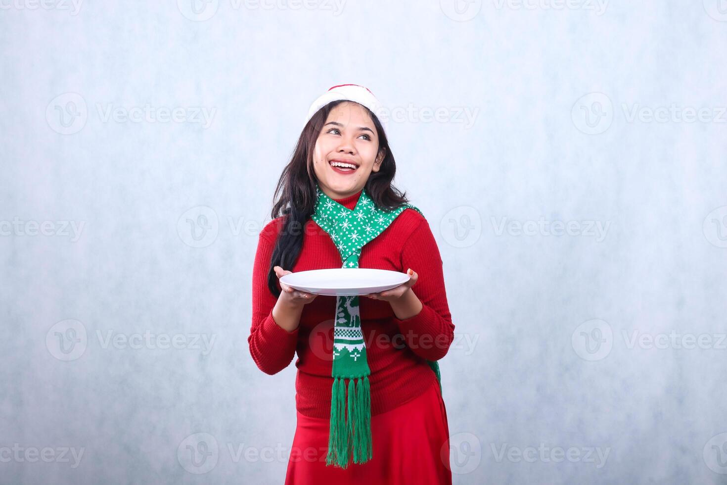 portrait of woman wearing red sweater red sweater with santa hat and scarf celebrating merry christmas and new year, cheerful candid over both hands carrying white plate, isolated on white background photo