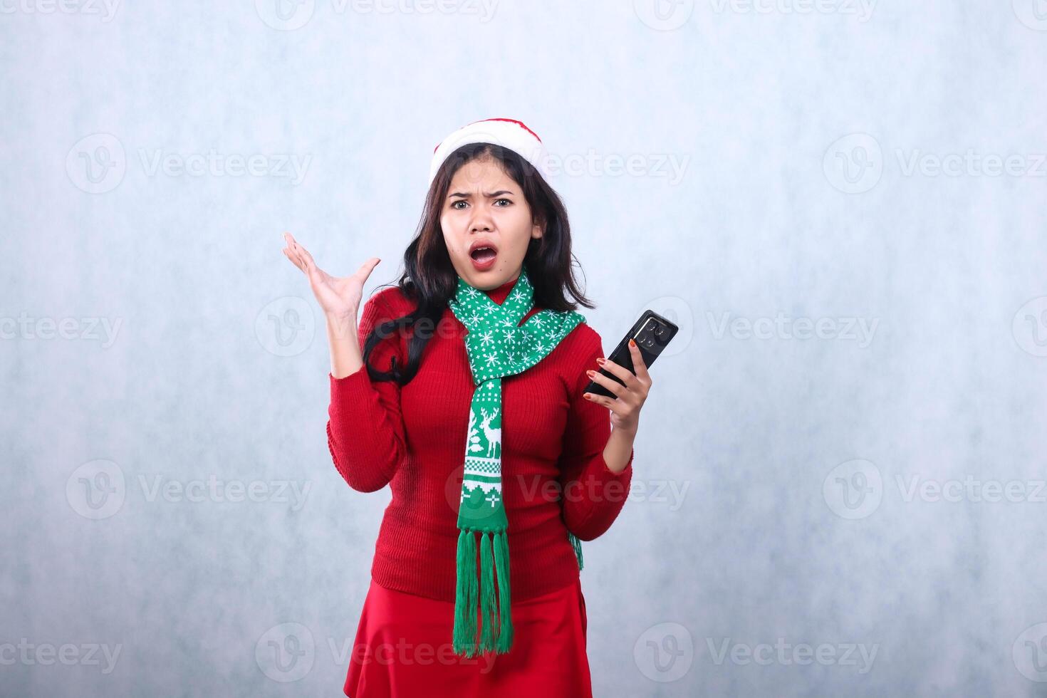 young asian woman surprised at camera holding mobile phone while growling,wearing red sweater with Santa hat and scarf celebrating merry christmas and new year,isolated on white background photo