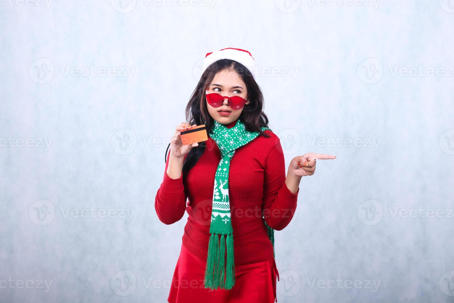 expression of woman wearing christmas sweater with santa hat and scarf, sarcastically looking to the left holding credit debit card, wearing glasses, pointing to the left, isolated on white background photo