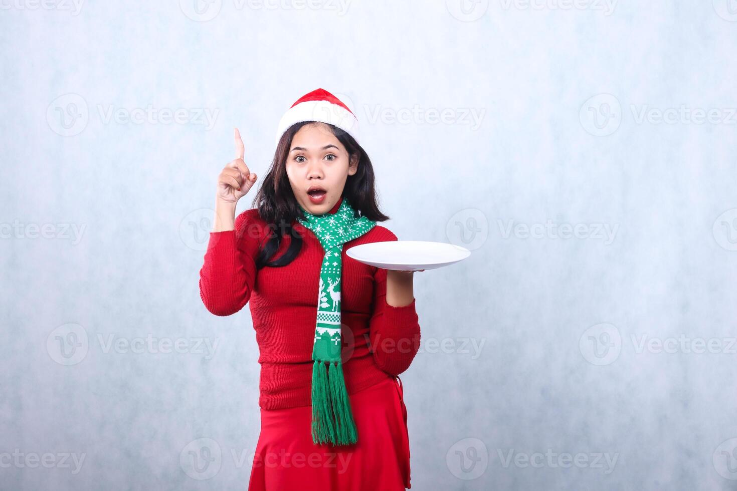 gesture of Asian woman wearing red sweater with Santa hat and scarf, surprised to camera holding white plate while pointing up, celebrating merry christmas and new year isolated on white background photo