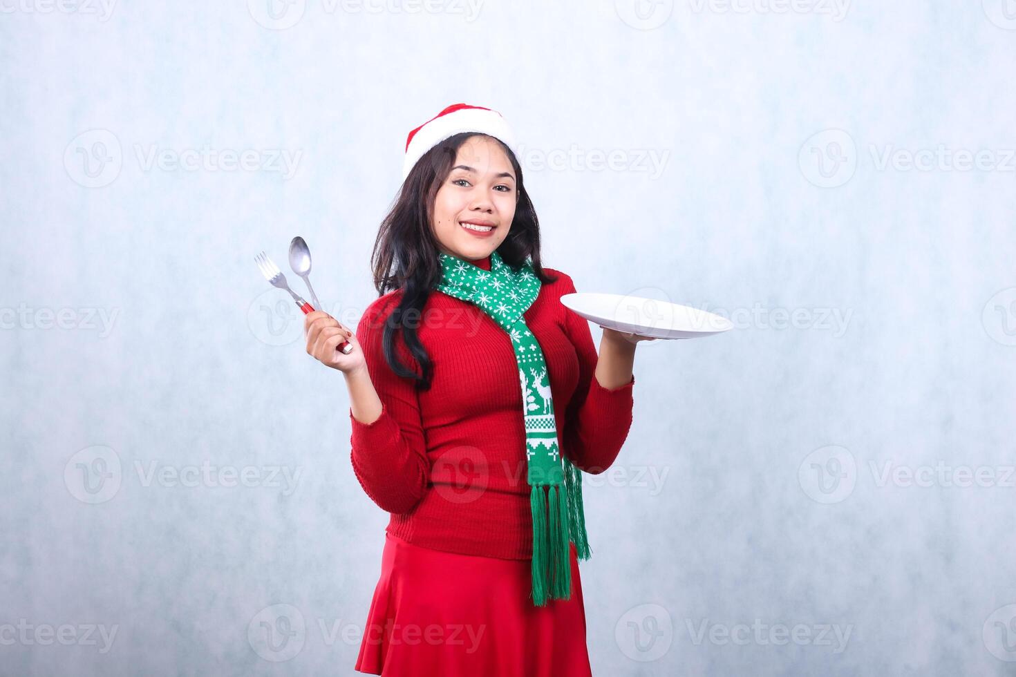 expression of indonesia woman wearing red sweater red sweater with Santa hat and scarf celebrating merry christmas, smile to camera carry white plate and hold spoon fork, isolated on white background photo