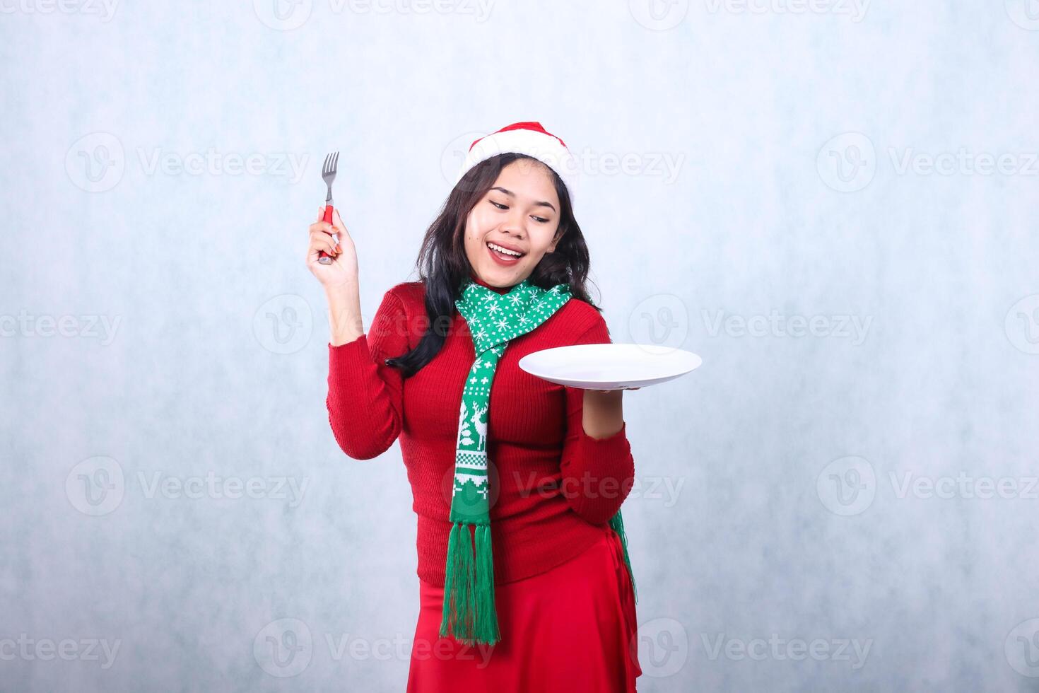 portrait adult woman wearing red sweater with Santa hat and scarf, cheerful to the plate carrying white plate and holding fork aside, celebrating merry christmas and new year isolated on white photo